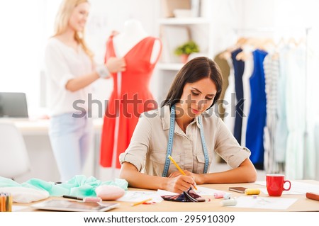 They like what they do. Confident young woman sketching while another women measuring dress on background