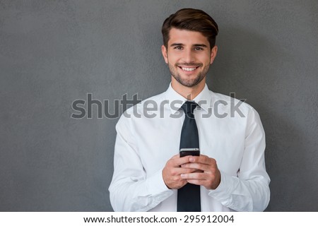 Staying in touch with my colleagues. Handsome young man in shirt and tie holing mobile phone and smiling while standing against grey background