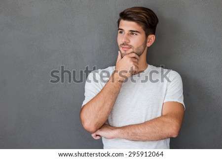 Lost in thoughts. Thoughtful young man holding hand on chin and looking away while standing against grey background