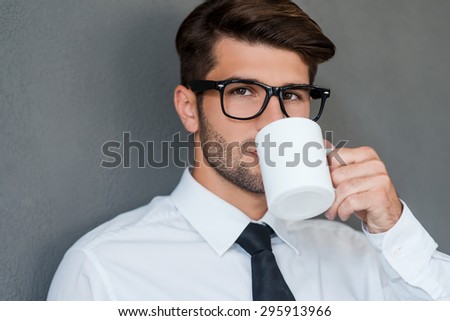 Inspired with cup of fresh coffee. Confident young man in shirt and tie drinking coffee and looking away while standing against grey background