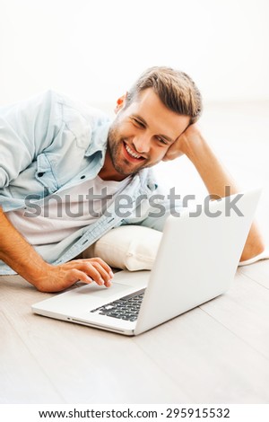 Comfortable place for working. Happy young man working on laptop and smiling while lying on the floor at his apartment