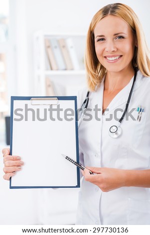 Your test results right here. Happy female doctor in white uniform looking at camera and smiling while pointing at clipboard