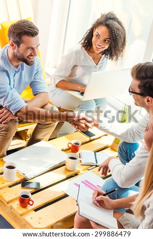 Welcome on board! Two cheerful young men sitting at the wooden desk in office and shaking hands while two beautiful women looking at them and smiling