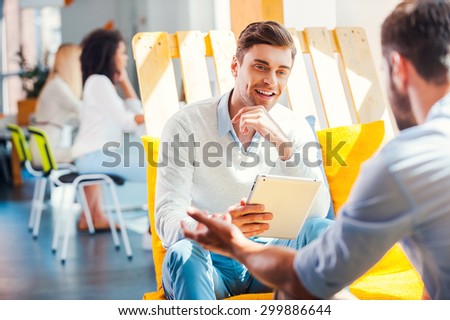 Collaboration is a key to success. Smiling young man holding digital tablet and discuss something with man sitting opposite him in the rest area of the office