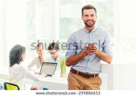 Enjoying office life. Joyful young man holding mobile phone and looking at camera while his colleagues working in the background