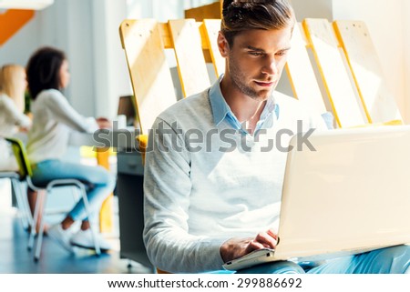 He is always hard at work. Concentrated young man working on laptop while his colleagues working in the background