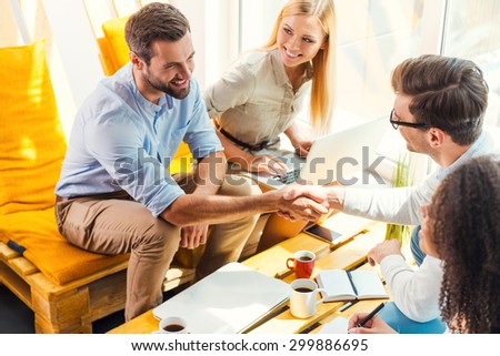 Congratulations! Two cheerful young men sitting at the wooden desk in office and shaking hands while two beautiful women looking at them and smiling