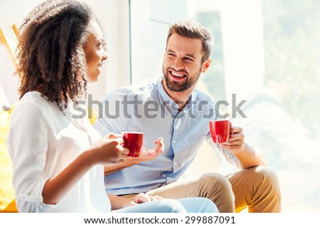 Sharing great ideas. Happy young African woman and young man holding cups of coffee and discussing something while sitting together near the window