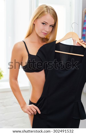 This dress will help me to impress! Smiling young woman in black bra and panties holding dress and looking at camera while standing indoors