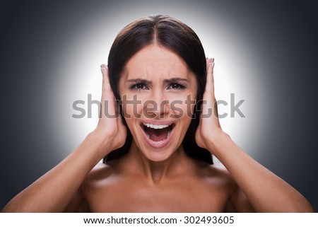 Too loud sound! Portrait of frustrated young shirtless woman shouting and covering ears by hands while standing against grey background