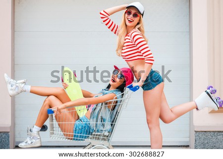 Having crazy day together. Side view of happy young woman on roller skates carrying her female friend in shopping cart and smiling while skating against the garage door