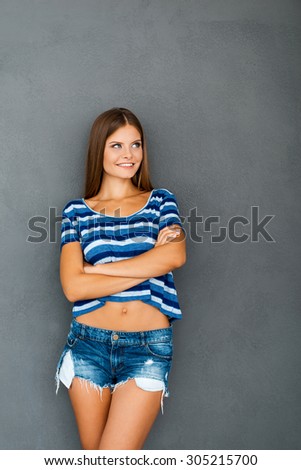 Cool and cute. Happy young woman keeping arms crossed and smiling while standing against grey background