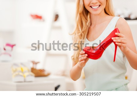 Perfect red heels. Cropped image of young woman holding red shoe and smiling while sitting in the shoe store