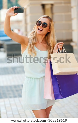 Shopping spree selfie. Joyful young woman holding shopping bags and making selfie on her smart phone while standing outdoors