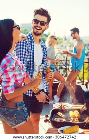 Perfect day for barbecue. Cheerful young couple barbecuing and holding bottles with beer while two people talking to each other in the background