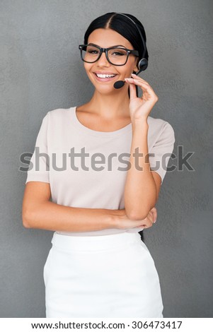 Always ready to help you. Smiling young female customer service looking at camera while standing against grey background