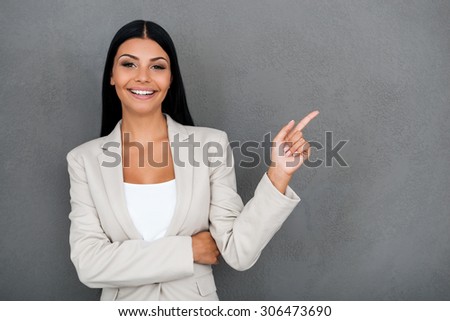 Beautiful and reliable. Smiling young businesswoman looking at camera and pointing away while standing against grey background