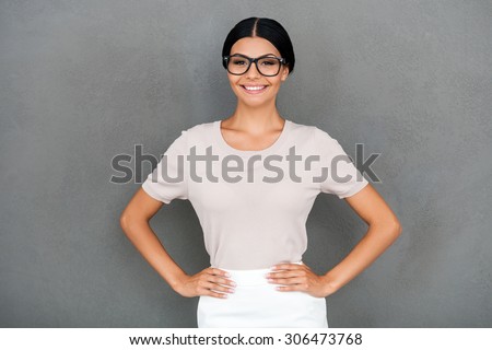 Proud of being herself. Smiling young businesswoman holding hands on hips and looking at camera while standing against grey background