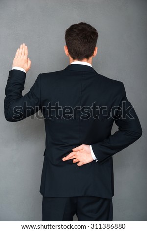 Telling lies. Rear view of young businessman keeping his fingers crossed and arm raised while standing against grey background