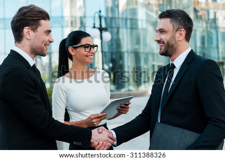 Glad to see you! Two happy young businessmen shaking hands while their female colleague holding digital tablet and smiling