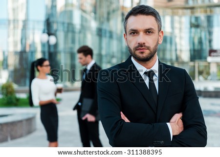 He is great team leader. Confident young businessman keeping arms crossed and looking at camera while two his colleagues talking to each other in the background