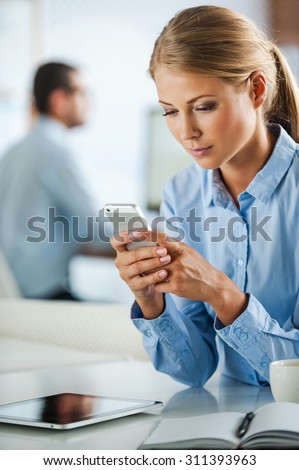 Staying in touch. Serious young businesswoman in formalwear holding mobile phone while sitting at her working place