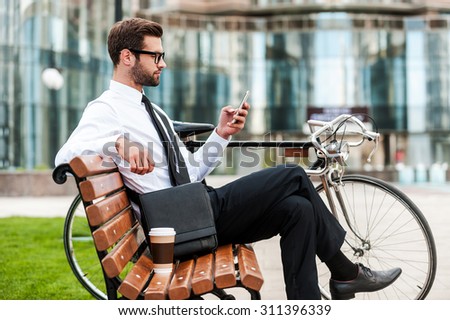 Young and successful. Side view of handsome young businessman holding mobile phone while sitting on the bench near his bicycle with office building in the background