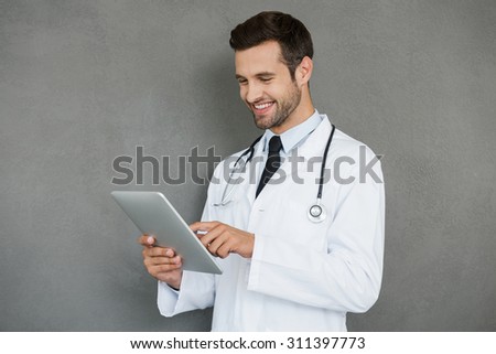 Digital solution for medical science. Smiling young doctor in white uniform working on digital tablet while standing against grey background