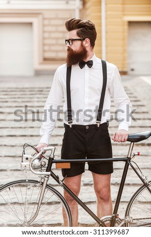 Man n style. Confident young bearded man holding hands on his bicycle and looking away while standing outdoors