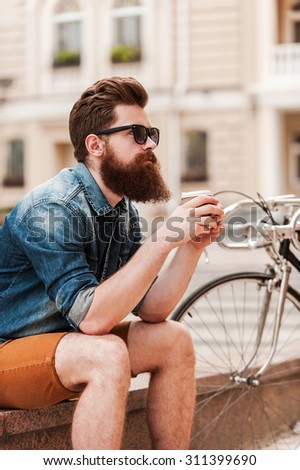Staying calm. Thoughtful young bearded man holding cup of coffee while sitting near his bicycle outdoors
