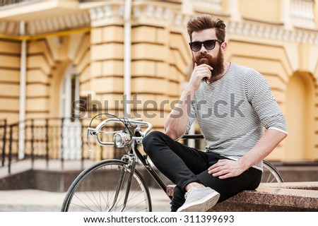Taking quick break. Thoughtful young bearded man holding hand on chin and looking at camera while sitting near his bicycle outdoors