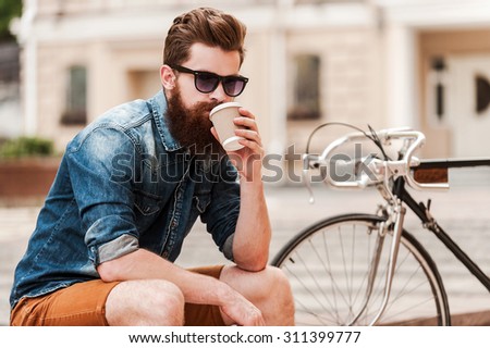 Enjoying fresh coffee. Confident young bearded man drinking coffee and looking at camera while sitting near his bicycle outdoors