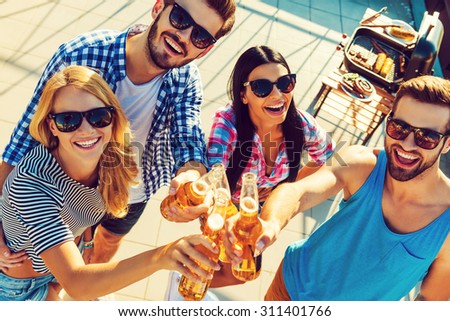 Celebrating their friendship. Top view of four cheerful young people clinking glasses with beer and looking at camera while standing outdoors