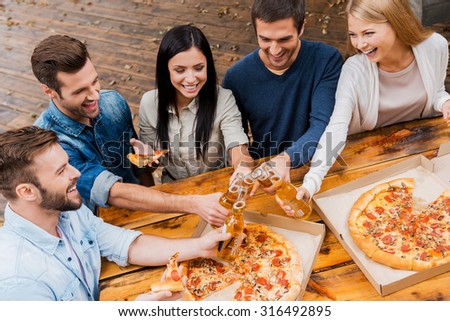 Finally it is Friday! Top view of five joyful young people clinking glasses with beer and eating pizza while standing outdoors
