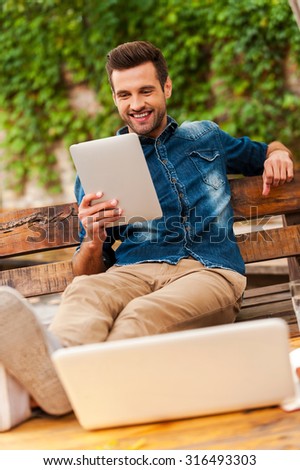 Technologies make life easier. Joyful young man holding digital tablet and smiling while sitting on the wooden bench outdoors