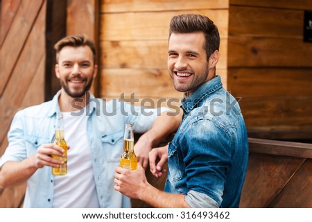 Old friends meeting. Two cheerful young men holding bottles with beer and looking at camera while leaning at the bar counter