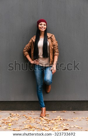 Beauty in style. Beautiful young woman leaning at the grey wall and smiling with fallen leaves laying around her
