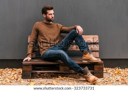 Casually handsome. Handsome young man sitting on the wooden pallet and looking away with grey wall in the background and orange fallen leaves on the floor