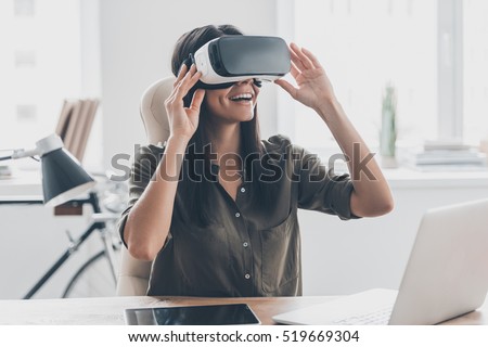 Future is right now. Confident young woman adjusting her virtual reality headset and smiling while sitting at her working place in office 商業照片 © 