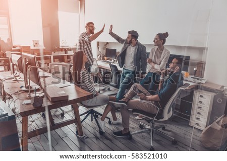Photo of We did it! Two cheerful young business people giving high-five while their colleagues looking at them and smiling 
