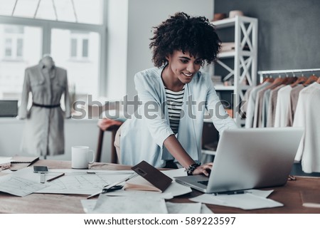 Photo of Responding on business e-mail. Beautiful young African woman working using computer and smiling while standing in workshop 