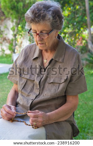 An old woman between 70 and 80 years old is praying in the morning in the garden sitting on the chair and keeping Anglican rosary in her hands.
