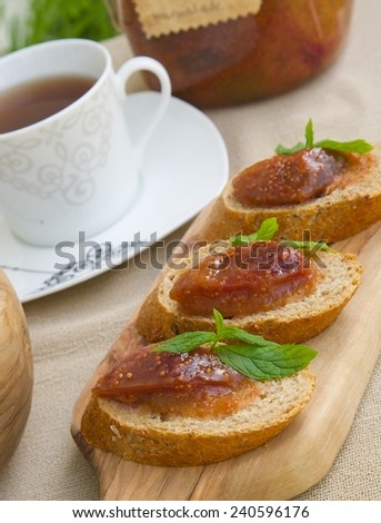 Pieces of bread with homemade fig marmalade on the wooden cutting board and a cup of tea. A glass with fig marmalade in the background