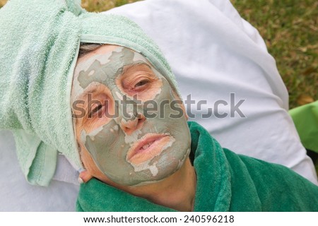 A senior woman between 70 and 80 years old is going through rejuvenating facial procedures. Table with spa skin care products in the background. Woman's face is covered by green clay mask. Close up