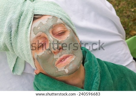 A senior woman between 70 and 80 years old is going through rejuvenating facial procedures. Table with spa skin care products in the background. Woman\'s face is covered by green clay mask