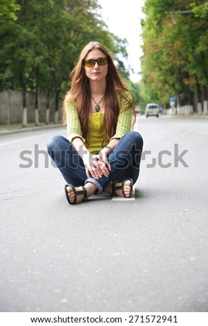A young girl between 20 and 30 years old in sun eyeglasses is sitting in the middle of the road. Free space for a text