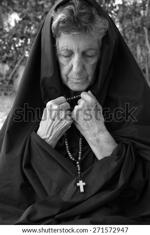 A senior woman between 70 and 80 years old, dressed in black, is praying in the garden. Black and white