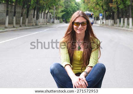A young girl between 20 and 30 years old in sun eyeglasses is sitting in the middle of the road. Free space for a text