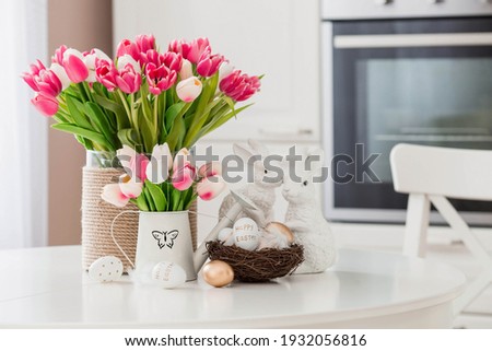 A bouquet of tulips, Easter bunnies and eggs with a golden pattern on the table. In the background is a white Scandinavian-style kitchen. Beautiful greeting card. The minimal concept.