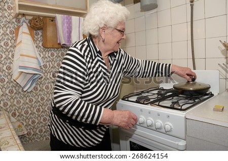 Senior woman prepares food on the gas stove in her kitchen. She takes cover of the frying pan.